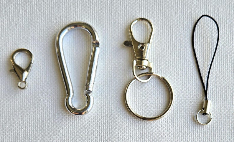 K&ouml;nnte beinhalten: Eine Auswahl an silberfarbenen Metallverschl&uuml;ssen und Zubeh&ouml;r. Dazu geh&ouml;ren ein kleiner Karabinerverschluss, ein Karabinerhaken, ein Drehverschluss mit Schl&uuml;sselring und ein schwarzes Schl&uuml;sselband mit kleinem Ring. Diese werden wahrscheinlich zum Befestigen von Gegenst&auml;nden verwendet.