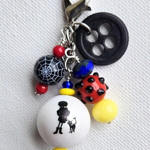 K&ouml;nnte beinhalten: Ein handgefertigter Charm mit einem silbernen Verschluss, mit einem schwarzen Knopf, einer Spinnennetzperle und verschiedenen bunten Perlen. Eine wei&szlig;e Perle mit einer Silhouette einer Person und einer Katze ist ebenfalls enthalten.