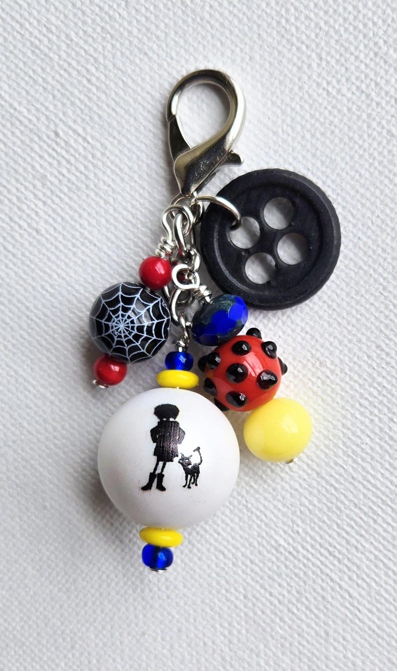 K&ouml;nnte beinhalten: Ein handgefertigter Charm mit einem silbernen Verschluss, mit einem schwarzen Knopf, einer Spinnennetzperle und verschiedenen bunten Perlen. Eine wei&szlig;e Perle mit einer Silhouette einer Person und einer Katze ist ebenfalls enthalten.