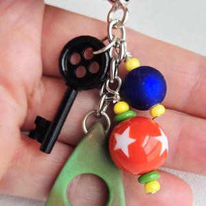 May include: A keychain with a black key charm, a green triangle charm, and colorful beads. The beads include a blue sphere, a red sphere with white stars, and yellow and green accents. The keychain is held by a silver heart-shaped clasp.