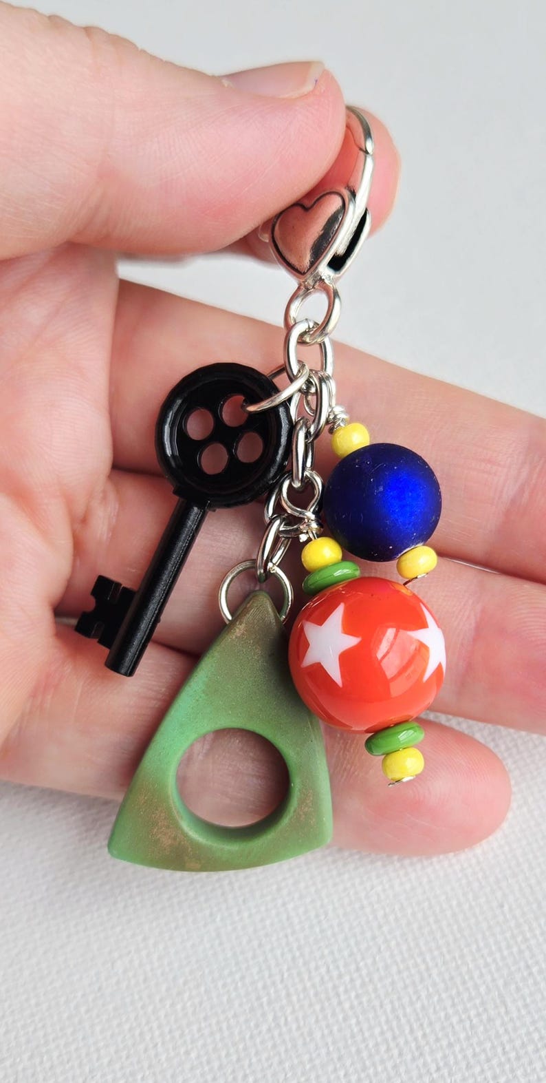 May include: A keychain with a black key charm, a green triangle charm, and colorful beads. The beads include a blue sphere, a red sphere with white stars, and yellow and green accents. The keychain is held by a silver heart-shaped clasp.