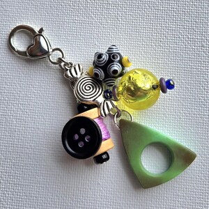 K&ouml;nnte beinhalten: Ein handgefertigter Charm-Anh&auml;nger mit einem herzf&ouml;rmigen Verschluss. Der Charm-Anh&auml;nger hat eine schwarz-wei&szlig;e Perle, eine gelbe Glasperle, ein gr&uuml;nes Dreieck, einen schwarzen Knopf und andere dekorative Elemente. Der Charm ist an einer silberfarbenen Kette befestigt.