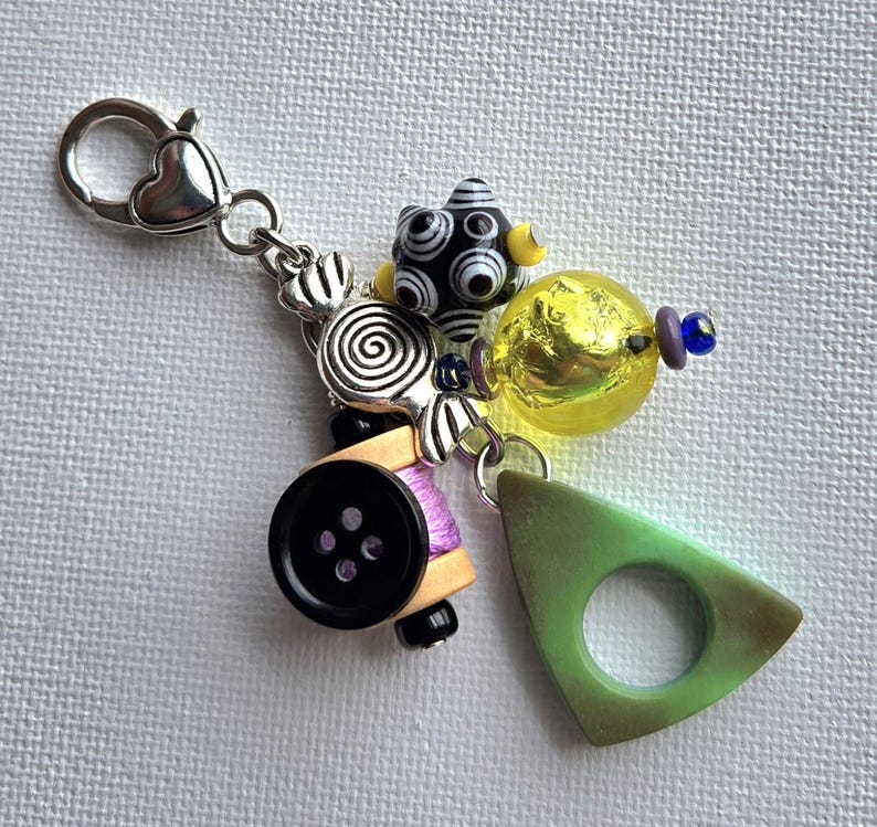 K&ouml;nnte beinhalten: Ein handgefertigter Charm-Anh&auml;nger mit einem herzf&ouml;rmigen Verschluss. Der Charm-Anh&auml;nger hat eine schwarz-wei&szlig;e Perle, eine gelbe Glasperle, ein gr&uuml;nes Dreieck, einen schwarzen Knopf und andere dekorative Elemente. Der Charm ist an einer silberfarbenen Kette befestigt.