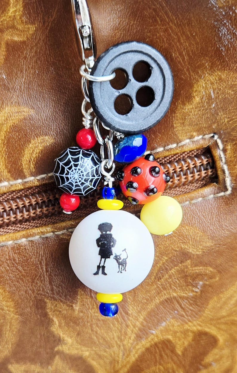 K&ouml;nnte beinhalten: Ein handgefertigter Schl&uuml;sselanh&auml;nger mit einem schwarzen Knopf, verschiedenen bunten Perlen und einem Anh&auml;nger mit der Silhouette einer Person mit einer Katze. Die Perlen umfassen Rot, Blau, Gelb und ein Spinnennetz-Design.