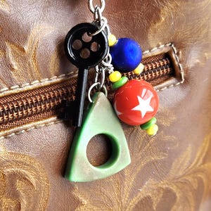 May include: A silver keychain with a heart-shaped clasp holding a black key, a green triangle with a hole, and colorful beads. A red ball with a white star is also attached. The background is a brown leather surface.