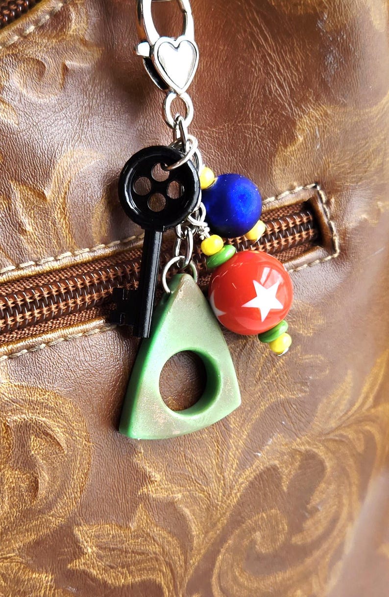 May include: A silver keychain with a heart-shaped clasp holding a black key, a green triangle with a hole, and colorful beads. A red ball with a white star is also attached. The background is a brown leather surface.