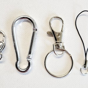 May include: Four silver-colored metal clasps and connectors of various designs. Includes a heart-shaped clasp, a carabiner-style clasp, a swivel clasp with a key ring, and a black lanyard with a small ring.