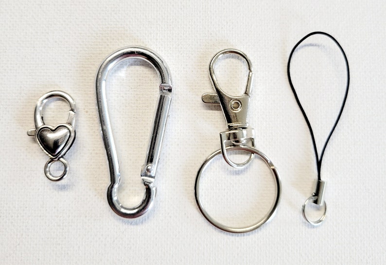 May include: Four silver-colored metal clasps and connectors of various designs. Includes a heart-shaped clasp, a carabiner-style clasp, a swivel clasp with a key ring, and a black lanyard with a small ring.