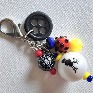 K&ouml;nnte beinhalten: Ein dekorativer Charm mit einem schwarzen Knopf, einem silbernen Verschluss und einer Sammlung farbiger Perlen. Die Perlen umfassen eine rote Perle im Marienk&auml;fer-Stil, eine schwarze Perle mit einem wei&szlig;en Spinnennetz-Design und eine wei&szlig;e Perle mit einer schwarzen Silhouette.