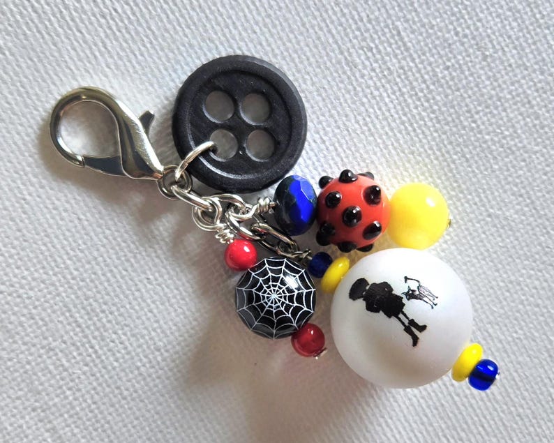 K&ouml;nnte beinhalten: Ein dekorativer Charm mit einem schwarzen Knopf, einem silbernen Verschluss und einer Sammlung farbiger Perlen. Die Perlen umfassen eine rote Perle im Marienk&auml;fer-Stil, eine schwarze Perle mit einem wei&szlig;en Spinnennetz-Design und eine wei&szlig;e Perle mit einer schwarzen Silhouette.