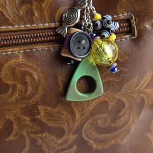 K&ouml;nnte beinhalten: Eine braune Ledertasche mit dekorativem Rei&szlig;verschluss und einem silbernen Schl&uuml;sselanh&auml;nger. Der Schl&uuml;sselanh&auml;nger hat verschiedene Anh&auml;nger, darunter einen Knopf, eine Garnrolle und bunte Perlen. Ein gr&uuml;ner Dreiecksanh&auml;nger ist ebenfalls befestigt.