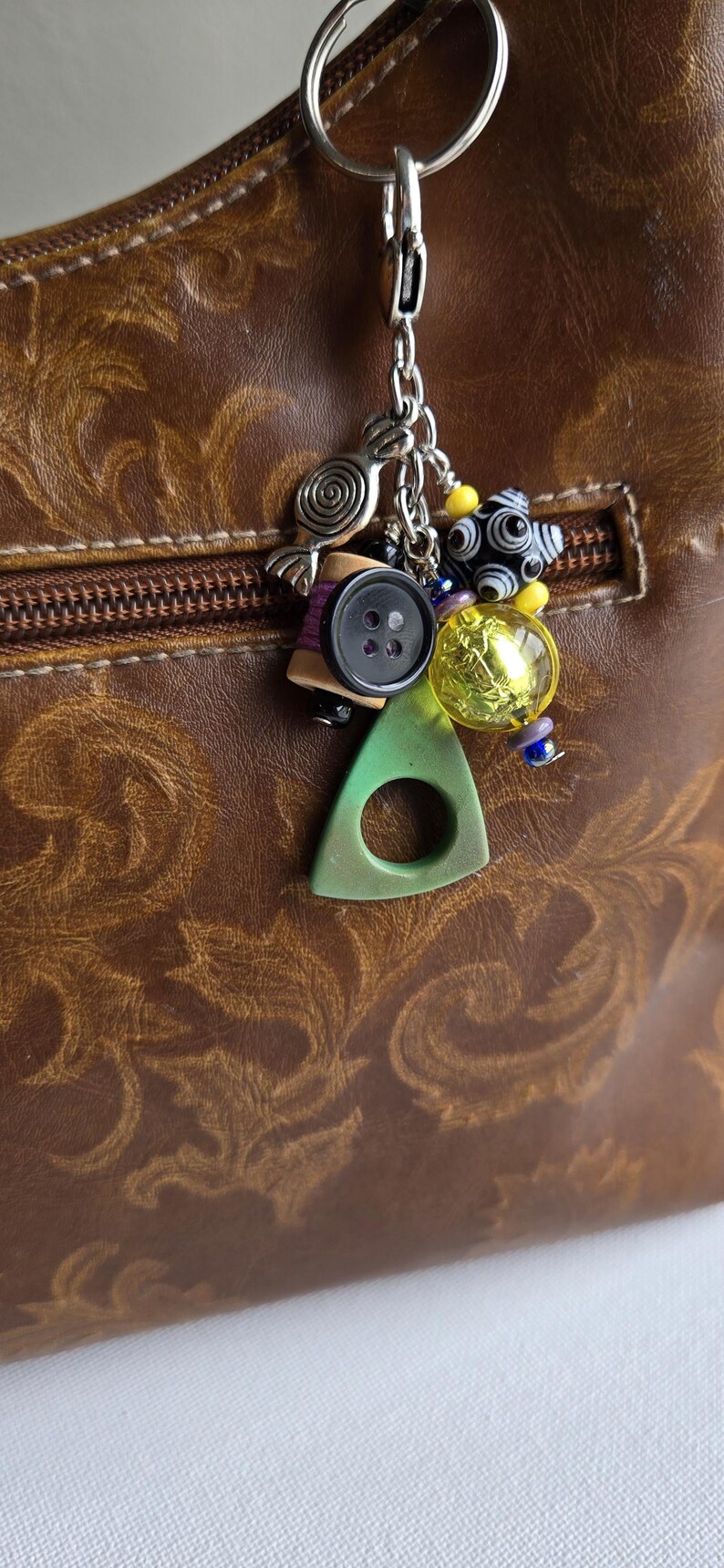 K&ouml;nnte beinhalten: Eine braune Ledertasche mit dekorativem Rei&szlig;verschluss und einem silbernen Schl&uuml;sselanh&auml;nger. Der Schl&uuml;sselanh&auml;nger hat verschiedene Anh&auml;nger, darunter einen Knopf, eine Garnrolle und bunte Perlen. Ein gr&uuml;ner Dreiecksanh&auml;nger ist ebenfalls befestigt.