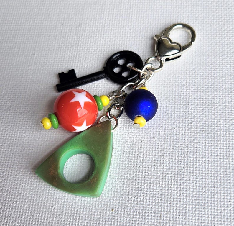 May include: A decorative keychain with a silver heart-shaped clasp. It features a black key charm, a red and white star bead, a blue bead, and a green triangle charm with a hole. The beads are accented with yellow and green.