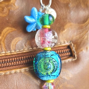 May include: A decorative bag charm featuring a silver-toned heart-shaped clasp, a light blue starfish charm, and a vertical arrangement of glass beads. The beads include a green cylinder, a clear sphere with glitter, and a large blue-green ammonite bead.