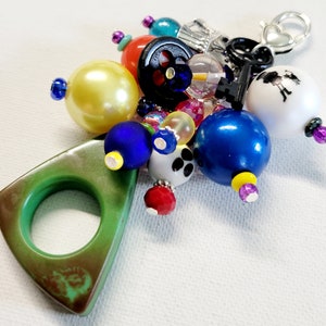 Pu&ograve; includere: Un ciondolo triangolare verde con un anello, circondato da perline colorate, tra cui blu, giallo, rosso, bianco e nero. Il ciondolo &egrave; attaccato a una chiave d'argento con un ciondolo a forma di cuore.