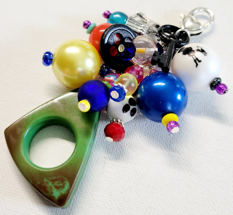 Pu&ograve; includere: Un ciondolo triangolare verde con un anello, circondato da perline colorate, tra cui blu, giallo, rosso, bianco e nero. Il ciondolo &egrave; attaccato a una chiave d'argento con un ciondolo a forma di cuore.