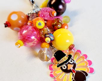 May include: A colorful charm with a pink, orange, and yellow color scheme. The charm features a small turkey with a black hat and a brown body. The charm is attached to a silver clip.