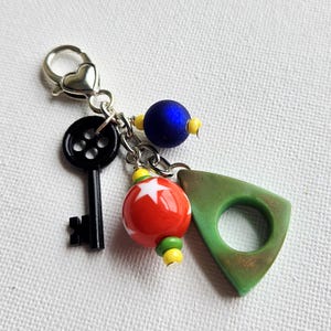 May include: A handmade keychain with a silver heart-shaped clasp. The keychain features a black key, a red and white star-patterned bead, a blue bead, and a green triangle-shaped charm. The beads are accented with yellow and green beads.