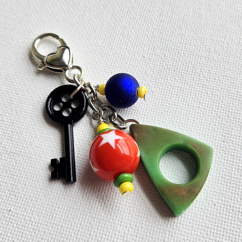 May include: A handmade keychain with a silver heart-shaped clasp. The keychain features a black key, a red and white star-patterned bead, a blue bead, and a green triangle-shaped charm. The beads are accented with yellow and green beads.