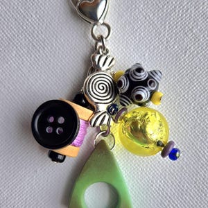 K&ouml;nnte beinhalten: Ein Charm mit verschiedenen Perlen und Anh&auml;ngern. Es enth&auml;lt einen schwarzen Knopf, eine silberne Bonbonspirale, ein gr&uuml;nes Dreieck mit einem Loch, eine gelbe Perle und eine schwarz-wei&szlig;e sternf&ouml;rmige Perle. Der Charm ist an einem silbernen Verschluss befestigt.