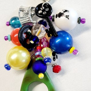 Pu&ograve; includere: Un ciondolo verde con un anello al centro, attaccato a una chiusura argentata con un ciondolo a forma di cuore. Il ciondolo &egrave; circondato da una variet&agrave; di perline colorate, tra cui blu, gialle, rosse, bianche e nere.