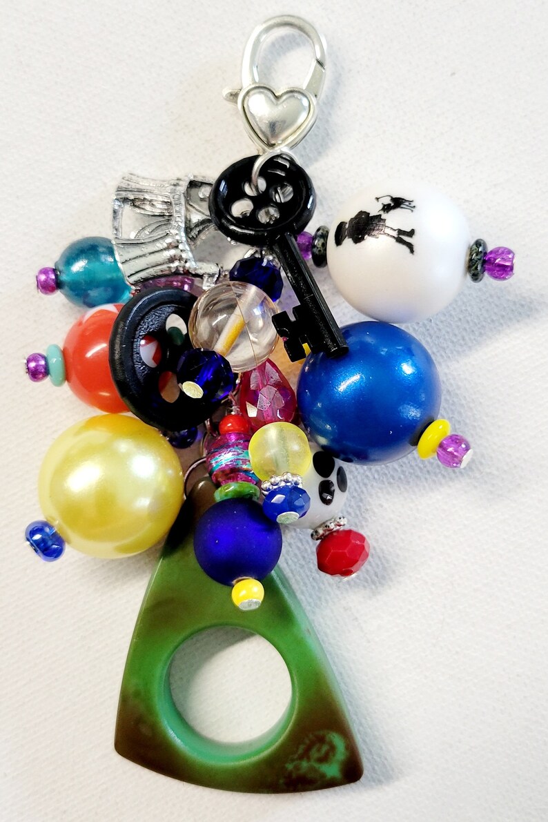 Pu&ograve; includere: Un ciondolo verde con un anello al centro, attaccato a una chiusura argentata con un ciondolo a forma di cuore. Il ciondolo &egrave; circondato da una variet&agrave; di perline colorate, tra cui blu, gialle, rosse, bianche e nere.