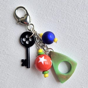 May include: A silver-toned keychain with a heart-shaped clasp. The keychain features a black key, a red and white star-patterned bead, a blue bead, and a green triangle charm with a hole.
