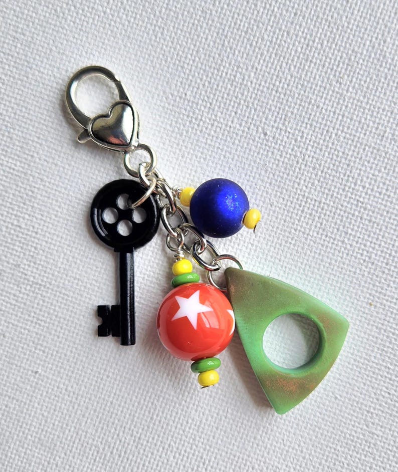 May include: A silver-toned keychain with a heart-shaped clasp. The keychain features a black key, a red and white star-patterned bead, a blue bead, and a green triangle charm with a hole.