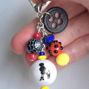 K&ouml;nnte beinhalten: Ein Charm mit einem silbernen Verschluss. Es zeigt einen schwarzen Knopf, verschiedene Perlen und eine wei&szlig;e Perle mit einer Silhouette einer Person und eines Hundes. Die Perlen umfassen ein Spinnennetz-Design, rote, blaue und gelbe Akzente.