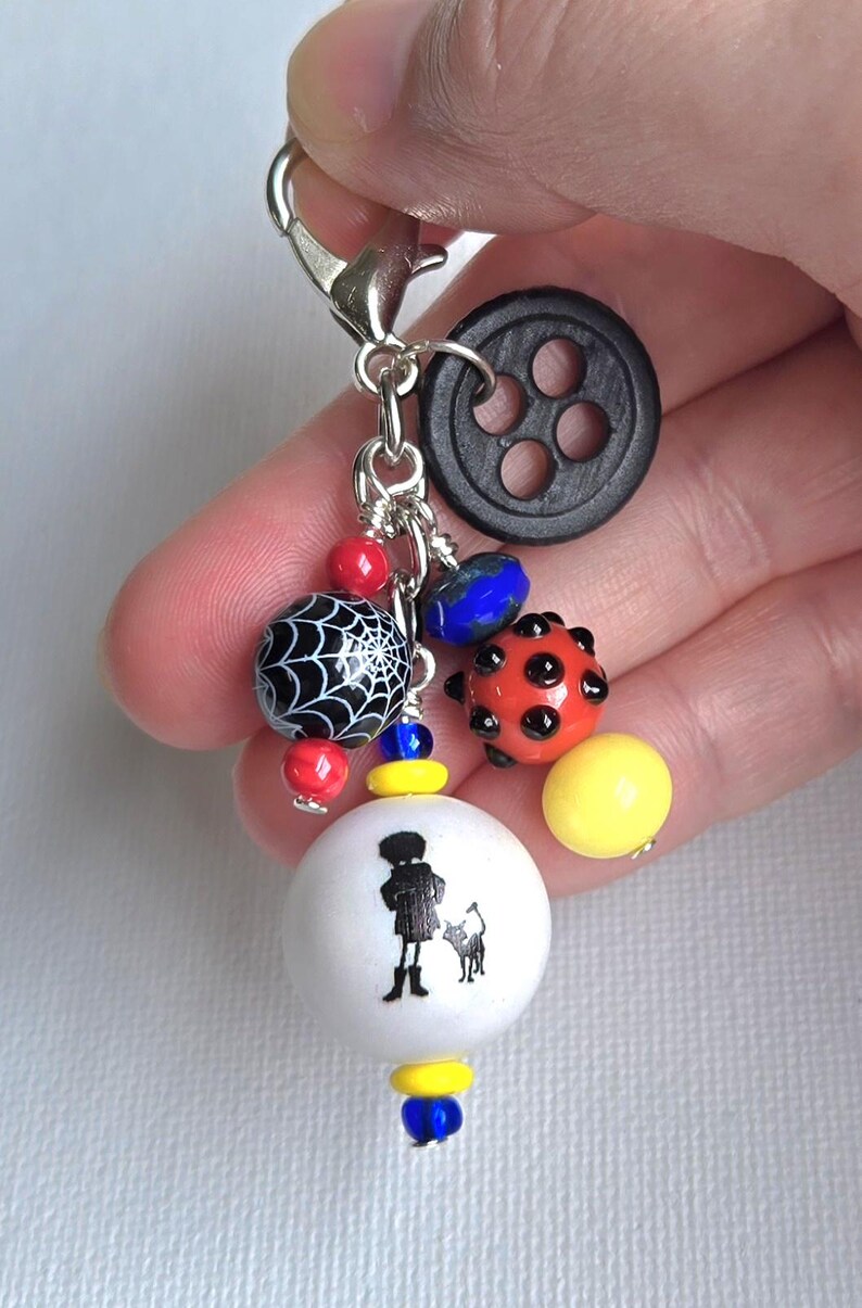 K&ouml;nnte beinhalten: Ein Charm mit einem silbernen Verschluss. Es zeigt einen schwarzen Knopf, verschiedene Perlen und eine wei&szlig;e Perle mit einer Silhouette einer Person und eines Hundes. Die Perlen umfassen ein Spinnennetz-Design, rote, blaue und gelbe Akzente.
