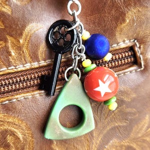 May include: A silver keychain with a heart-shaped clasp, featuring a black key, a green triangle with a hole, and colorful beads. A red bead with a white star is also included.