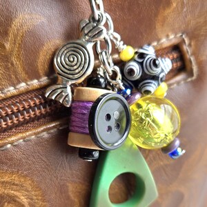 K&ouml;nnte beinhalten: Eine Sammlung von Charms an einer silbernen Kette, darunter eine Spirale, eine Spule mit lila Faden, ein schwarzer Knopf, ein gr&uuml;nes Dreieck und verschiedene Perlen. Die Charms sind an einer braunen Lederoberfl&auml;che befestigt.