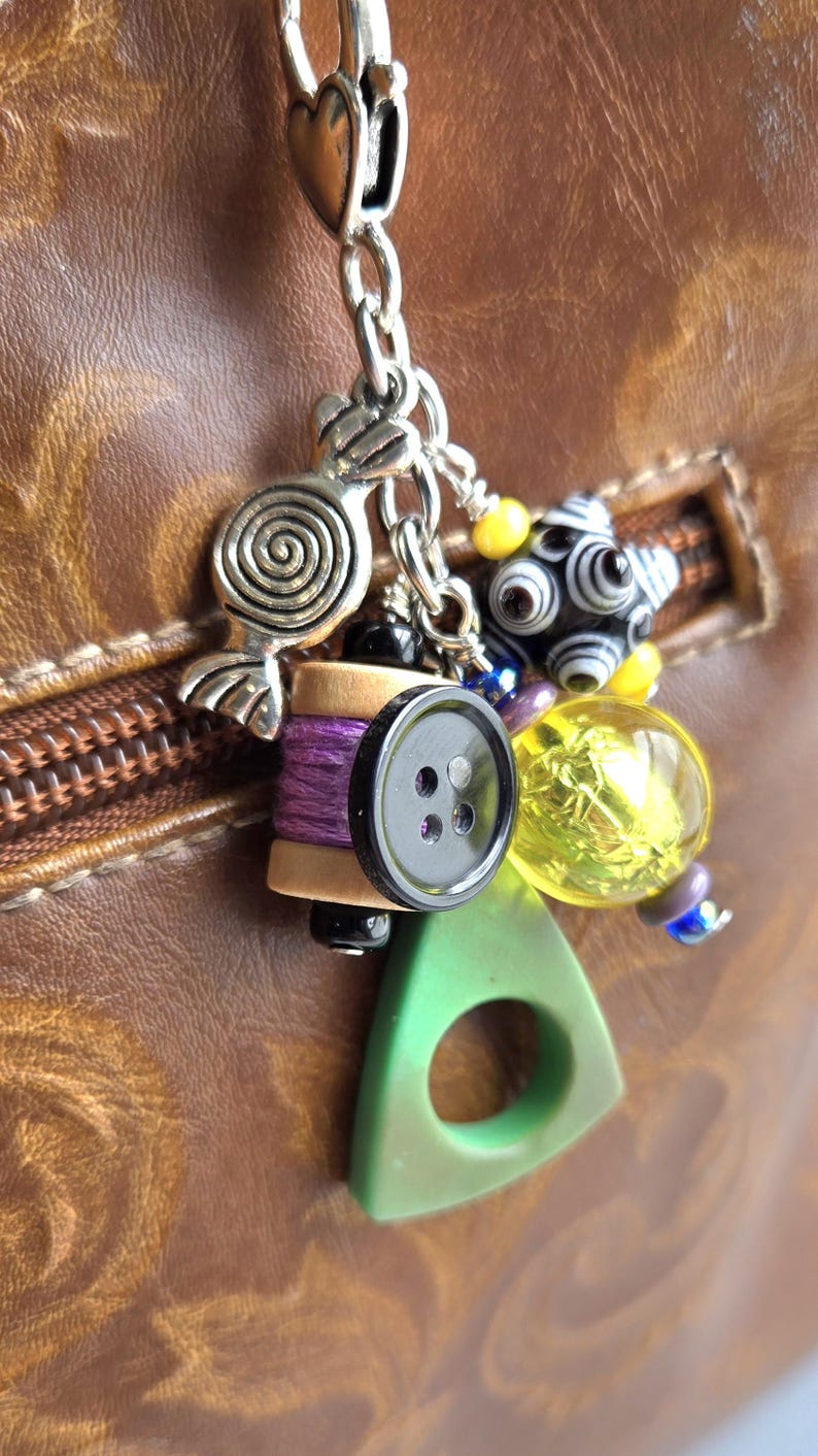 K&ouml;nnte beinhalten: Eine Sammlung von Charms an einer silbernen Kette, darunter eine Spirale, eine Spule mit lila Faden, ein schwarzer Knopf, ein gr&uuml;nes Dreieck und verschiedene Perlen. Die Charms sind an einer braunen Lederoberfl&auml;che befestigt.