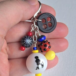 K&ouml;nnte beinhalten: Ein handgefertigter Charm-Anh&auml;nger mit einem schwarzen Knopf, verschiedenen Perlen und einer wei&szlig;en Kugel mit einer schwarzen Silhouette einer Person und einer Katze. Der Anh&auml;nger hat rote, blaue und gelbe Akzente und einen silberfarbenen Verschluss.