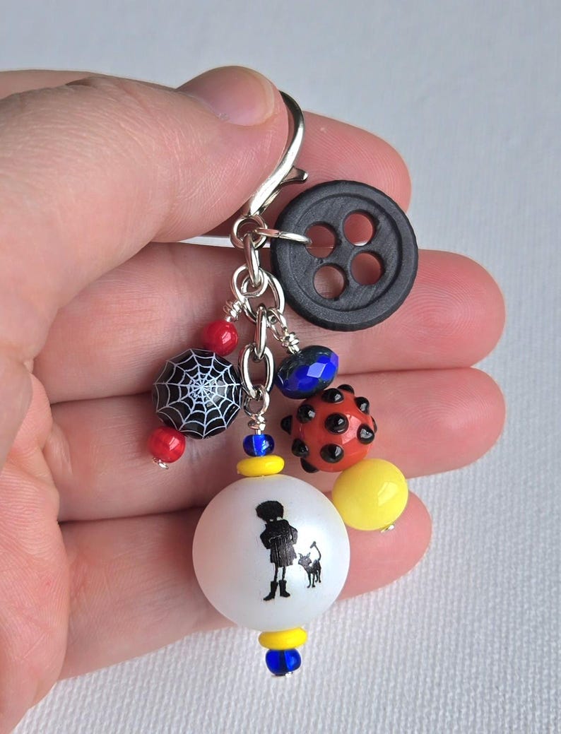 K&ouml;nnte beinhalten: Ein handgefertigter Charm-Anh&auml;nger mit einem schwarzen Knopf, verschiedenen Perlen und einer wei&szlig;en Kugel mit einer schwarzen Silhouette einer Person und einer Katze. Der Anh&auml;nger hat rote, blaue und gelbe Akzente und einen silberfarbenen Verschluss.