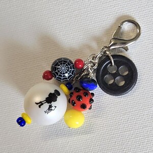 K&ouml;nnte beinhalten: Ein Charm mit verschiedenen Perlen und einem Verschluss. Der Charm enth&auml;lt einen schwarzen Knopf, eine Spinnennetzperle, rote und blaue Perlen, eine Marienk&auml;ferperle und eine wei&szlig;e Perle mit einem Silhouette-Design. Der Charm ist an einem silberfarbenen Verschluss befestigt.