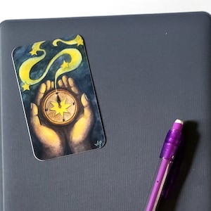 Könnte beinhalten: Ein graues Notizbuch mit einem Aufkleber, der eine Hand zeigt, die einen Kompass mit Sternen hält. Ein violetter Stift liegt auf dem Notizbuch.