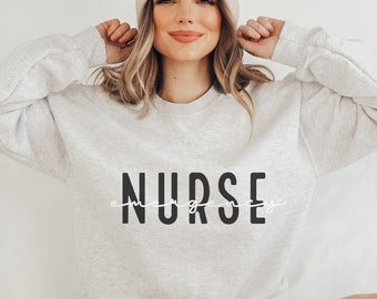 May include: A grey sweatshirt with the word "NURSE" printed in black letters with a white cursive "emergency" underneath.