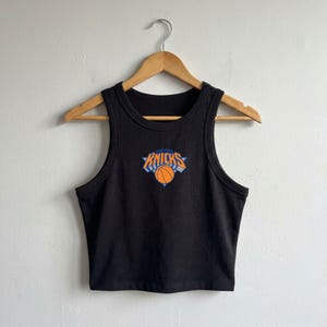 May include: A black sleeveless crop top featuring a centred graphic of the New York Knicks logo, which includes the text "NEW YORK KNICKS" in orange and blue lettering above an orange basketball illustration with blue accents on a white background.