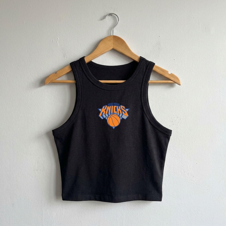 May include: A black sleeveless crop top featuring a centred graphic of the New York Knicks logo, which includes the text "NEW YORK KNICKS" in orange and blue lettering above an orange basketball illustration with blue accents on a white background.