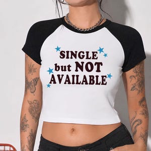 May include: A white short-sleeve raglan t-shirt with black sleeves and a crew neckline. The front features the dark brown text "SINGLE but NOT AVAILABLE" surrounded by small, glittery blue star graphics scattered across the chest area.