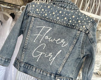 Könnte beinhalten: Hellblau gewaschene Jeansjacke für ein Blumenmädchen, verziert mit Kunstperlen entlang der Schultern. Auf der Rückseite der Jacke steht in weißer Schrift "Flower Girl". Die Jacke hat ein klassisches Jeansdesign mit Knopfverschlüssen.