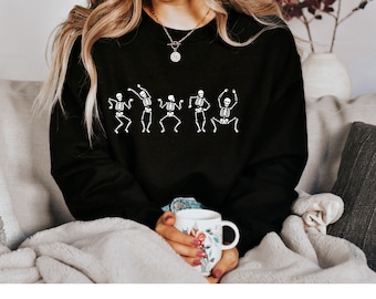 May include: Black sweatshirt with a white graphic of dancing skeletons. The sweatshirt is worn by a person sitting on a couch.