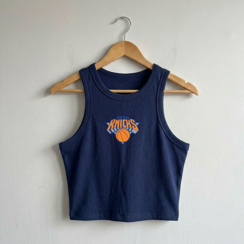 May include: A dark navy blue ribbed vest top featuring a central graphic of a basketball and the text "NEW YORK KNICKS" in orange and white. The garment is sleeveless with a round neckline and is displayed on a wooden hanger against a plain wall.