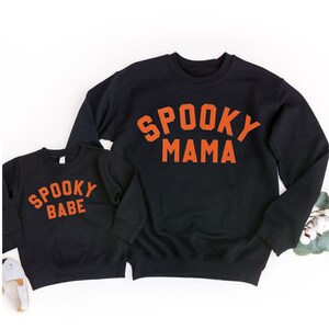 May include: Two black sweatshirts with orange text. The larger sweatshirt says "Spooky Mama" and the smaller sweatshirt says "Spooky Babe".