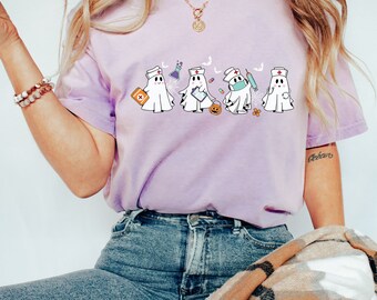 May include: A purple t-shirt featuring a design of four ghosts dressed as nurses. The ghosts are holding medical supplies such as a stethoscope, a syringe, a pill bottle, and a first aid kit. The ghosts are wearing white nurse hats and masks.