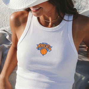 May include: A white ribbed tank top featuring the New York Knicks basketball team logo printed on the centre chest. The logo consists of the word Knicks in orange and blue lettering above an orange basketball graphic with a blue triangular outline.
