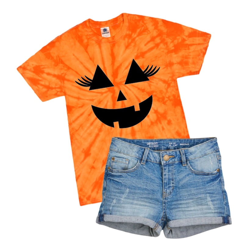 May include: Orange tie-dye t-shirt with a black pumpkin face graphic featuring eyelashes. The shirt is paired with blue denim shorts.