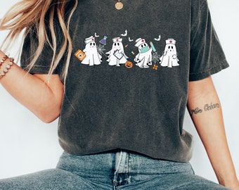 May include: A dark grey t-shirt featuring a graphic of four ghosts dressed as nurses. The ghosts are wearing surgical masks, holding medical supplies, and have a red cross on their hats. The graphic also includes a spiderweb, a pumpkin, and a bat.