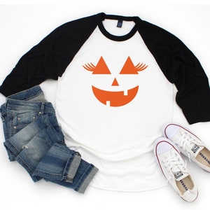 May include: A white and black baseball tee shirt with an orange jack-o'-lantern face graphic. The shirt is styled with blue jeans and white sneakers.